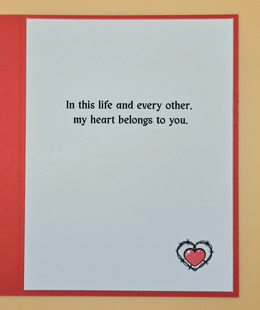 Card with romantic message 'In this life and every other, my heart belongs to you.' and a heart design on a beige background.