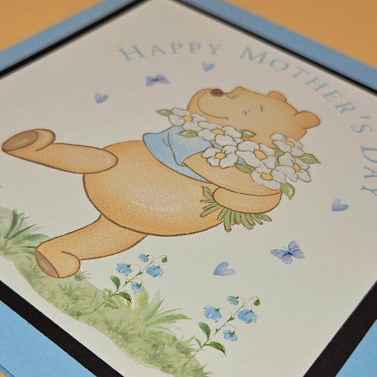Mother's Day card with a teddy bear holding flowers on a light blue border.