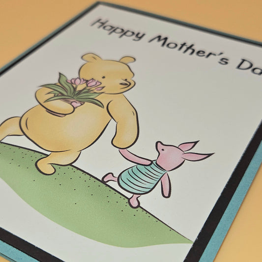Mother's Day card with a bear and piglet holding flowers on a grassy hill