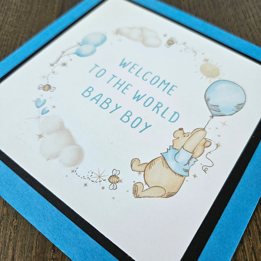 Baby shower card with Winnie the Pooh and text 'Welcome to the world baby boy' on a wooden surface.