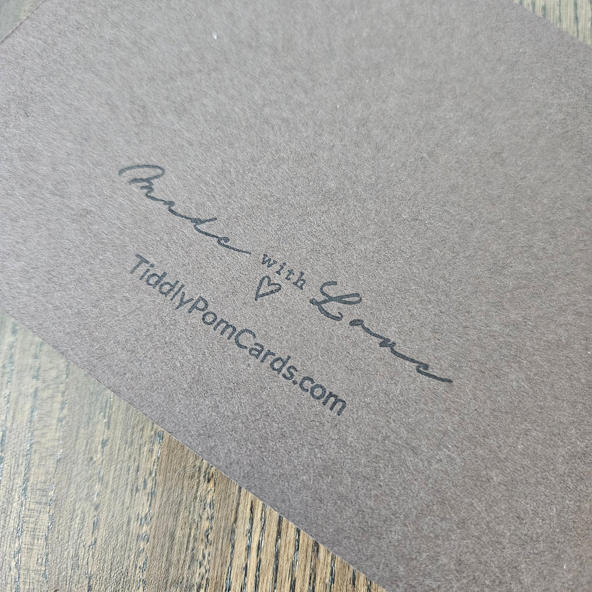 A close-up of a textured gray greeting card or packaging piece printed with the words 'Made with Love' and 'TiddlyPomCards.com' on a light wood surface.