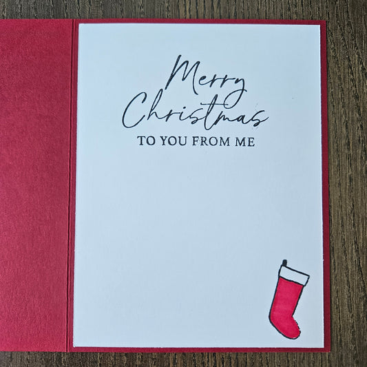 A square Christmas greeting card with a white front and red border, placed on a wooden surface. The card reads 'Merry Christmas TO YOU FROM ME' in black script and serif text, with a small red stocking illustration in the upper right corner.