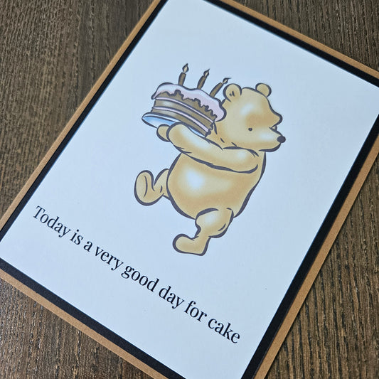Card with cartoon bear holding a cake and text 'Today is a very good day for cake' on a wooden surface.