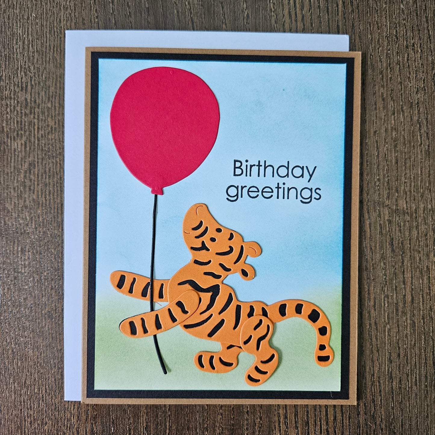 Handmade Winnie the Pooh birthday card featuring a die-cut illustration of Tigger holding a bright red balloon against a soft hand-inked watercolor sky and green meadow background.