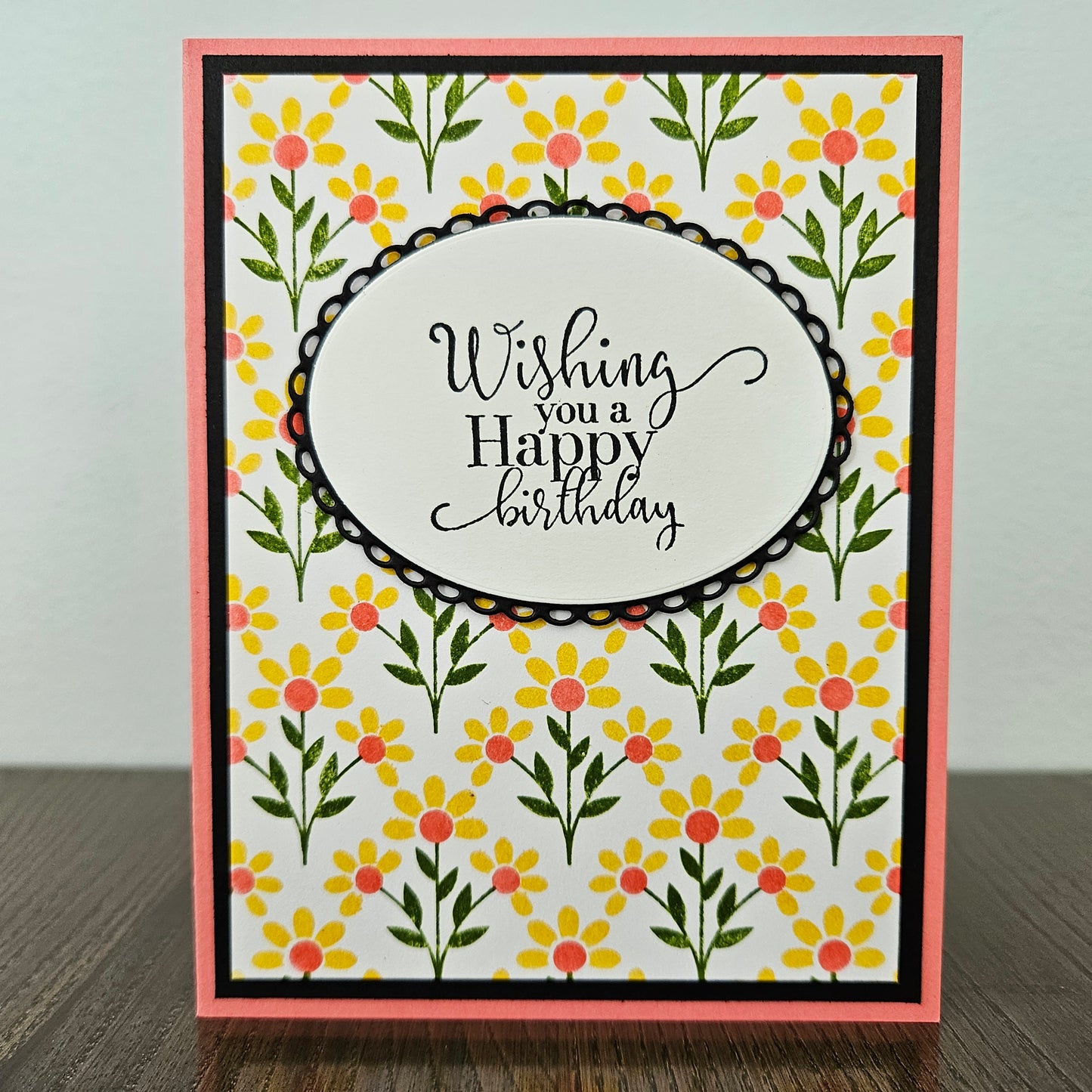 Handmade birthday card for her with hand-stamped yellow daisy field and coral centers with scalloped oval sentiment frame on black and pink border. Text reads Wishing you a Happy Birthday.