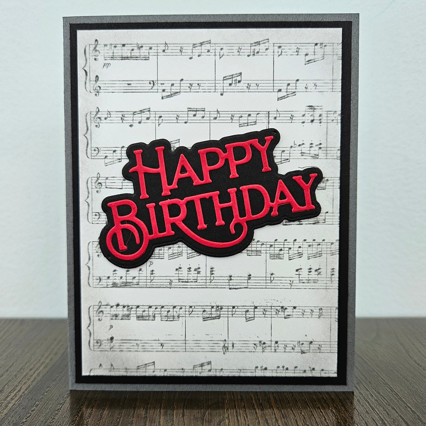 Handmade sheet music birthday card with vintage music score background and bold layered red and black die-cut Happy Birthday sentiment on black and grey border.