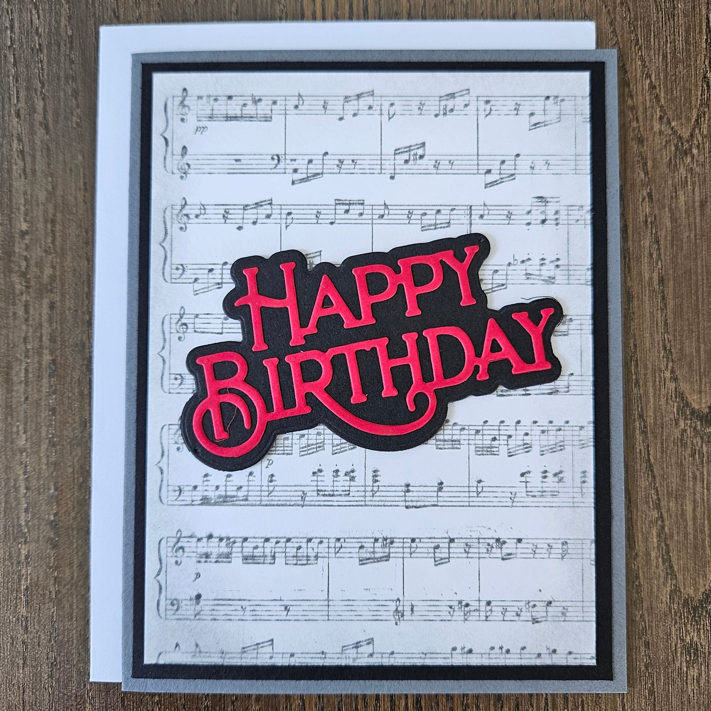 Handmade sheet music birthday card with vintage music score background and bold layered red and black die-cut Happy Birthday sentiment on black and grey border.