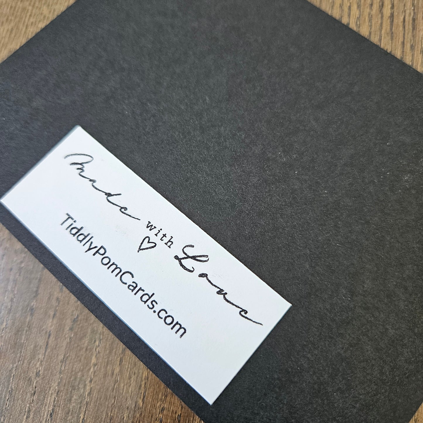 A close-up of a dark charcoal gray textured card or box cover with a white rectangular label that reads 'Made with Love' and 'TiddlyPomCards.com', placed on a wood surface.