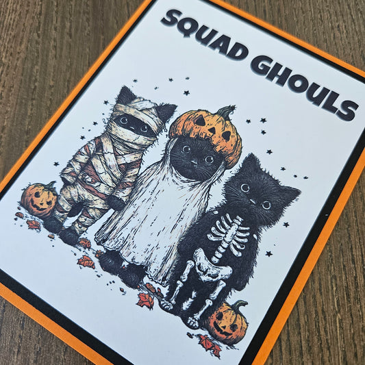 A square printed card or sticker featuring a Halloween illustration with the text 'SQUAD Ghouls' on a white background, bordered in orange and black. The artwork shows black cats dressed as skeletons, a mummy, and pumpkins on a wood surface.
