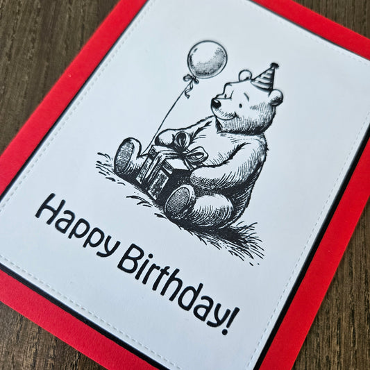 handmade A2 birthday card featuring a classic black ink storybook sketch illustration of Winnie the Pooh wearing a striped party hat while sitting with a wrapped gift box and a balloon, printed with the sentiment Happy Birthday!, finished with a stitched edge border, black mat, and red cardstock