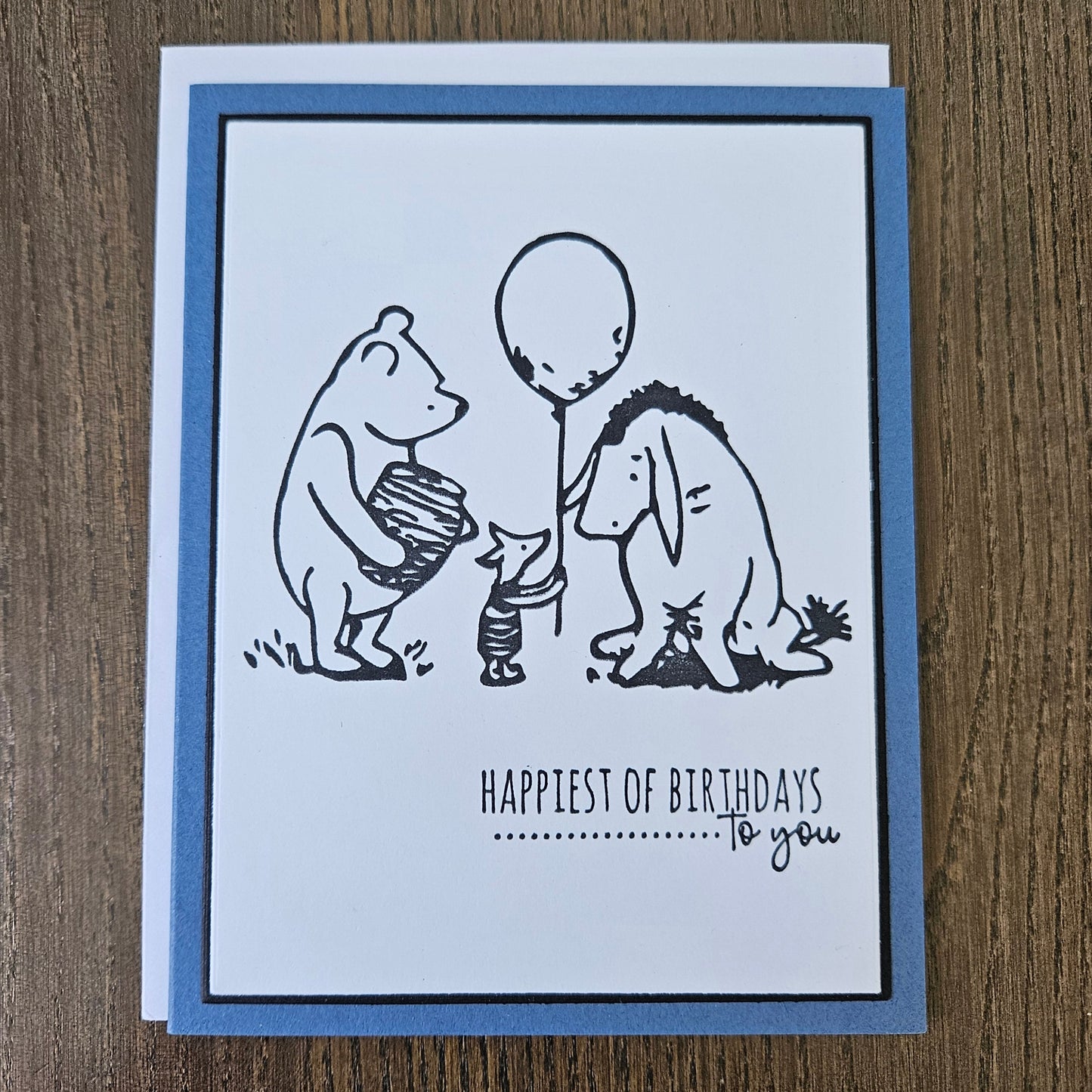 a handmade A2 birthday card featuring a classic black ink storybook sketch illustration of Winnie the Pooh with a honey pot, Piglet holding a balloon, and Eeyore standing together, printed with the sentiment Happiest of Birthdays To you, finished with a dark mat and blue cardstock frame