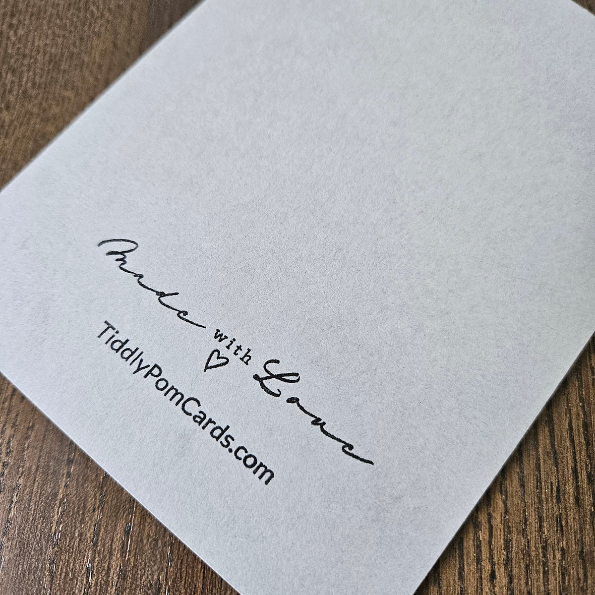 A close-up of a white textured paper pom card printed with the words 'Made with Love' and 'TidilyPomCards.com' in black script, resting on a brown wooden surface.