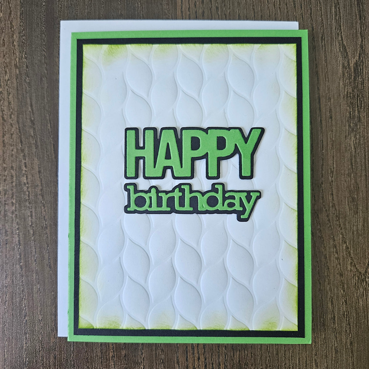 A handmade square greeting card with a white embossed wave pattern, featuring the words 'HAPPY birthday' in green and black. The card is framed with a green and black border and is photographed flat on a dark wood background.