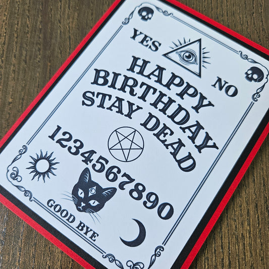 A gothic-style birthday card featuring a Ouija board design with the text 'YES NO HAPPY BIRTHDAY STAY DEAD 1234567890 GOOD BYE' along with illustrations of a cat, crescent moon, pentagram, eye, skulls, and sun, photographed on a wooden surface.