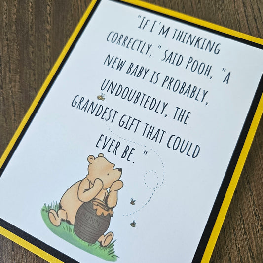 A square printed card or sign featuring a quote about Winnie the Pooh and a honey pot, photographed on a wooden surface. The card has a white center with black text, a yellow border, and a small illustration of Pooh holding a honey pot at the bottom left.