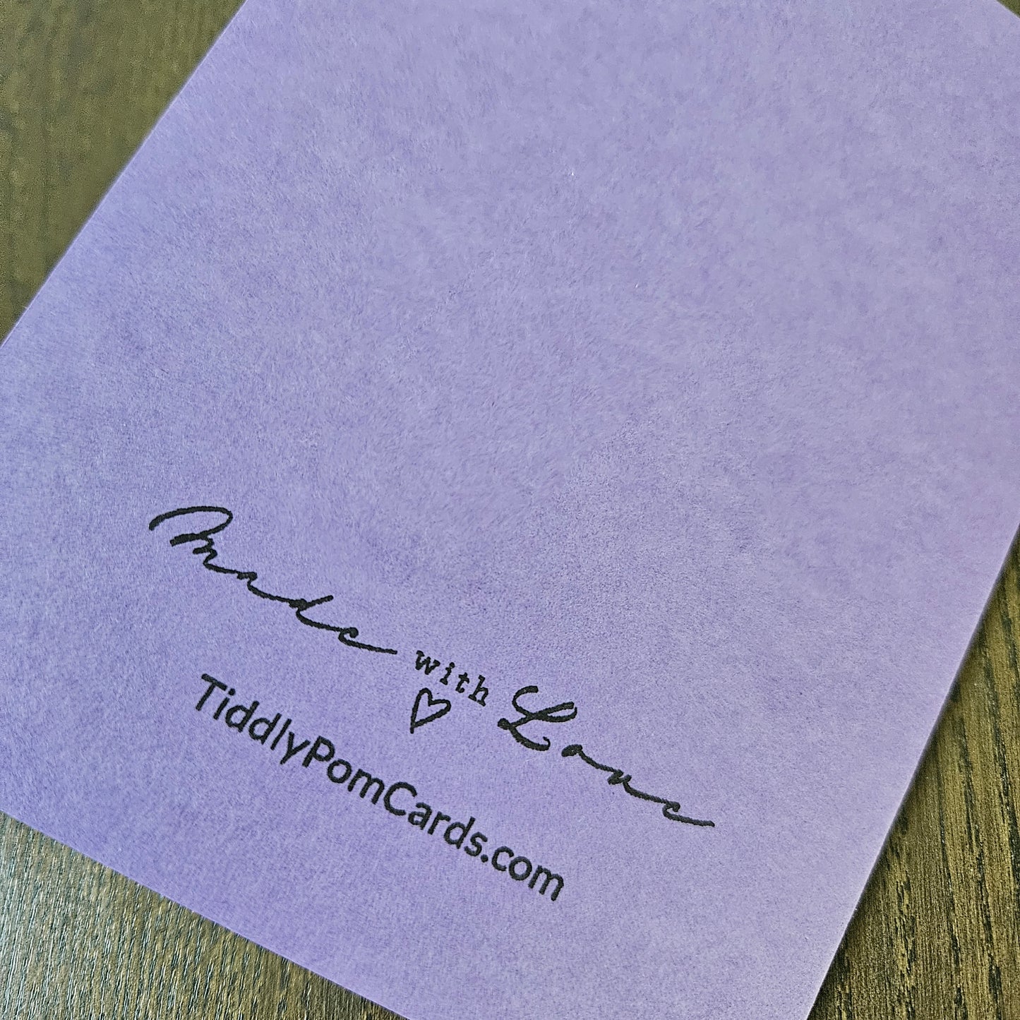 A close-up of a lavender purple textured card on a wooden surface, printed with the words 'Made with Love' and the website 'TiddlyPomCards.com'.