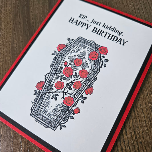 Front of a handmade A2 gothic birthday card featuring a layered design of red, black, and white cardstock. Above an ornate illustration of a coffin covered in red roses and vines is the snarky sentiment: RIP... just kidding, HAPPY BIRTHDAY.