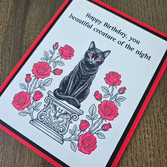 Front of a handmade A2 gothic birthday card featuring a black cat with red eyes sitting on a pedestal surrounded by red roses with the text Happy Birthday, you beautiful creature of the night.