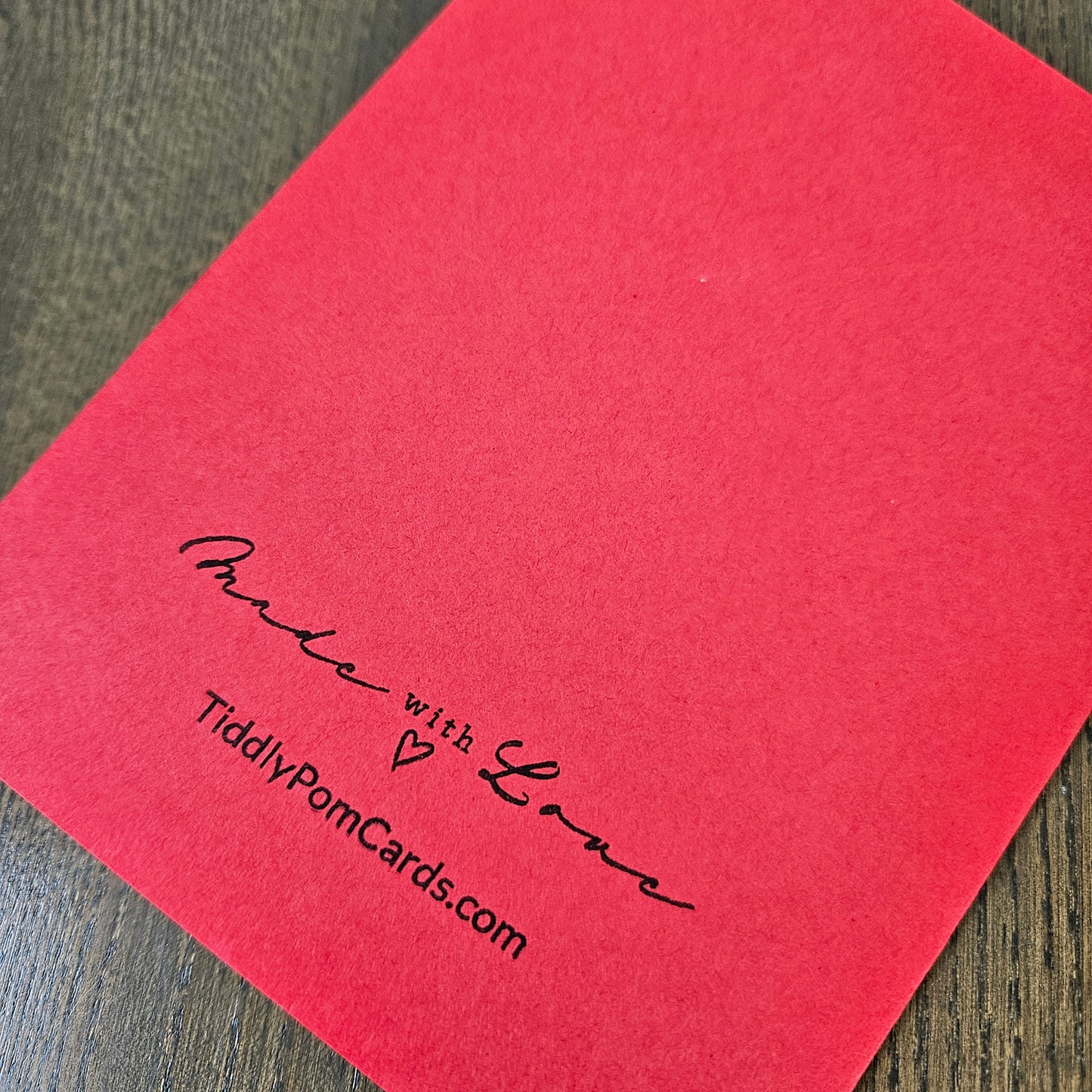 A red square pom card with black printed text reading 'Made with Love' and 'TidilyPomCards.com', placed on a dark wood surface.