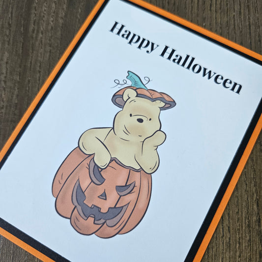 A square greeting card featuring Winnie the Pooh holding a carved pumpkin, with the text 'Happy Halloween' in black at the top left. The card has an orange border and is photographed on a dark wood surface.