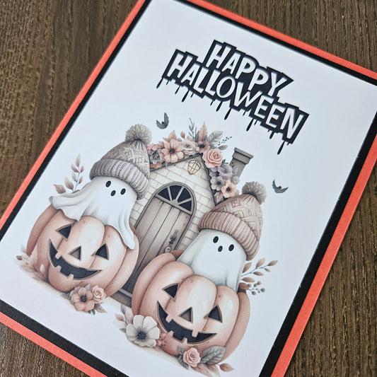 Front of a handmade A2 cute boho ghost Halloween card featuring layered orange and black cardstock framing two ghosts wearing beanie hats inside jack-o'-lantern pumpkins with text reading HAPPY HALLOWEEN.