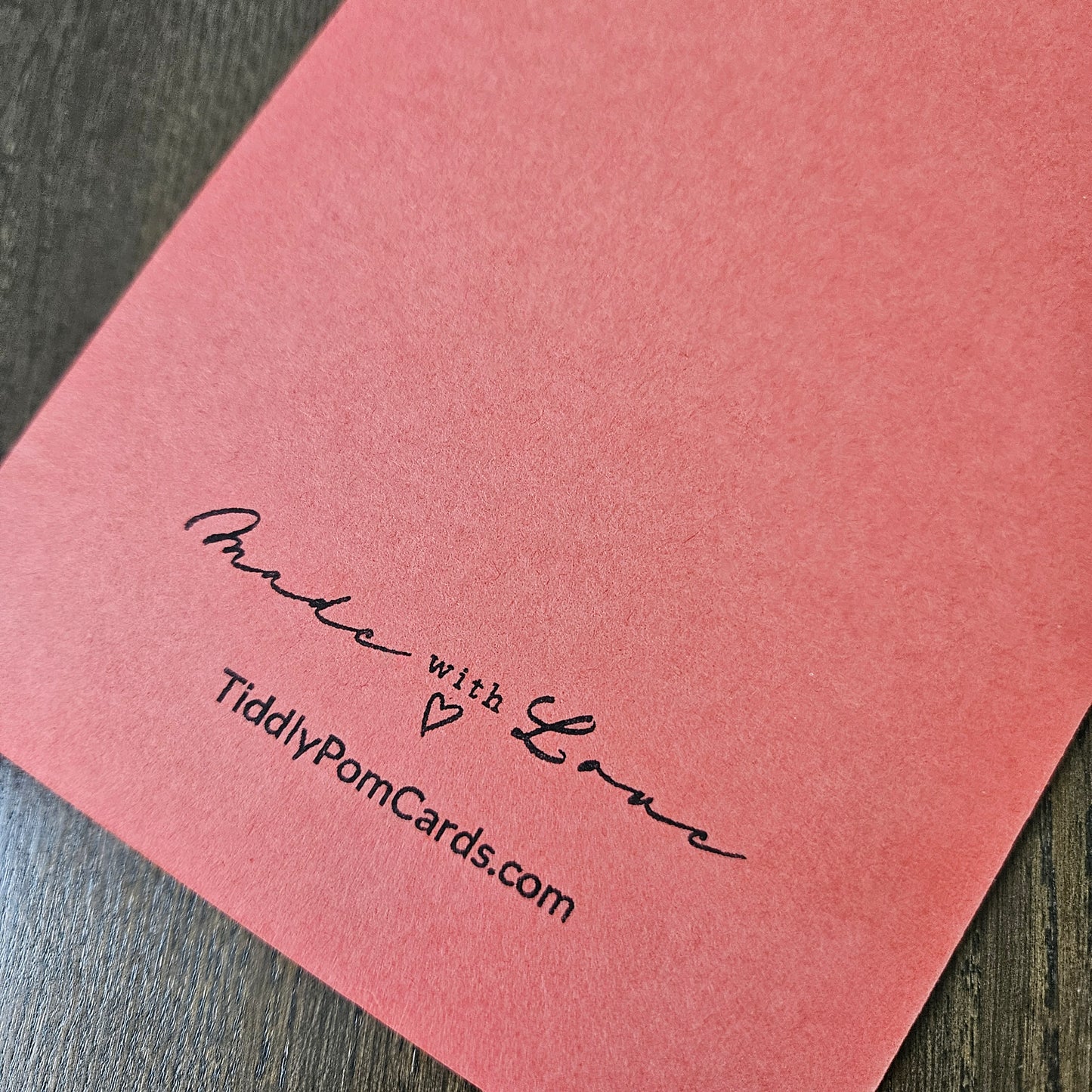 A close-up of a coral pink pom pom card featuring the text 'Made with Love' and 'TiddlyPomCards.com' printed in black script, resting on a dark wood surface.