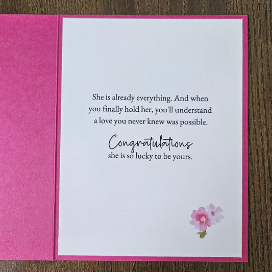 A square pink greeting card shown open on a wooden surface, with a white front insert card featuring black congratulatory text and a small pink flower illustration in the upper right.