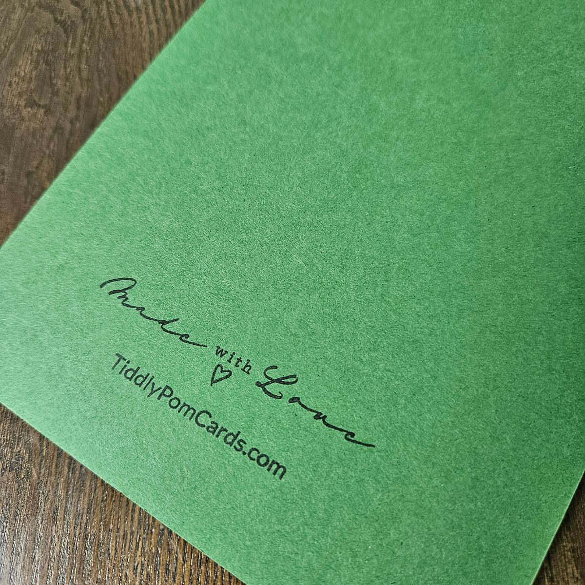 A green textured card or envelope corner with black printed script reading 'Praise with Love' and 'TidilyPomCards.com', photographed on a wooden surface.