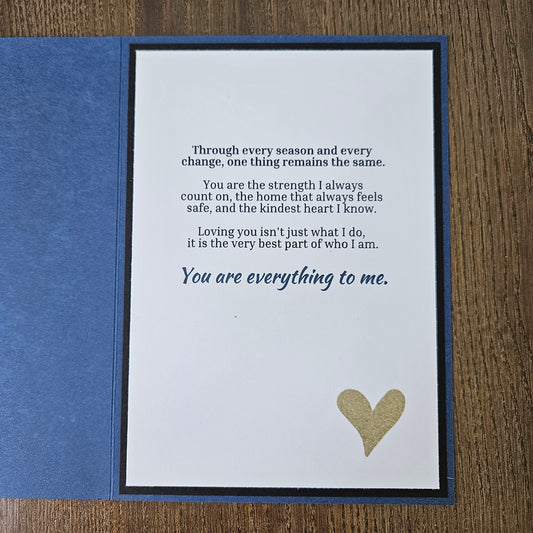 A square blue greeting card or gift card shown open on a wooden surface, with a white front insert featuring a gold heart and printed sentimental text, plus a blue backing piece.