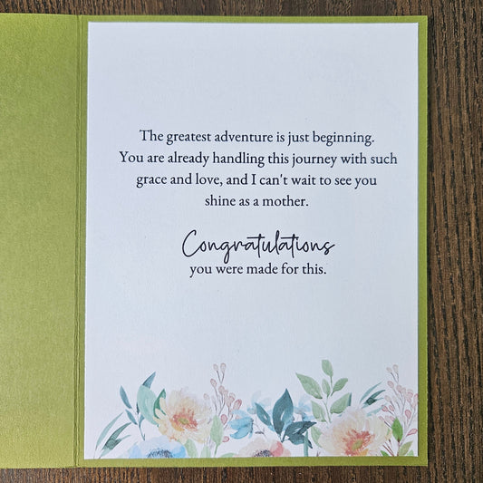 An open greeting card with a white front insert featuring a motivational quote and watercolor floral illustration at the bottom, set against an olive green backing on a dark wood surface.