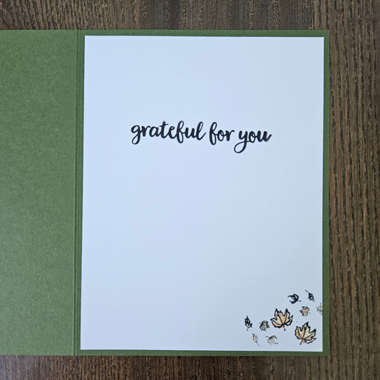A handmade greeting card featuring a white front insert with the words 'grateful for you' in black script, placed on a textured olive green backing. Small autumn leaves and birds are stamped in the bottom right corner.
