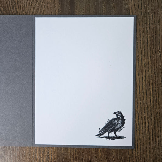 A square white art print or card with a dark gray border, featuring a black ink sketch of a raven near the bottom right corner. The print is displayed flat on a dark brown wood-grain surface, with a textured dark gray paper sheet partially visible on the left.