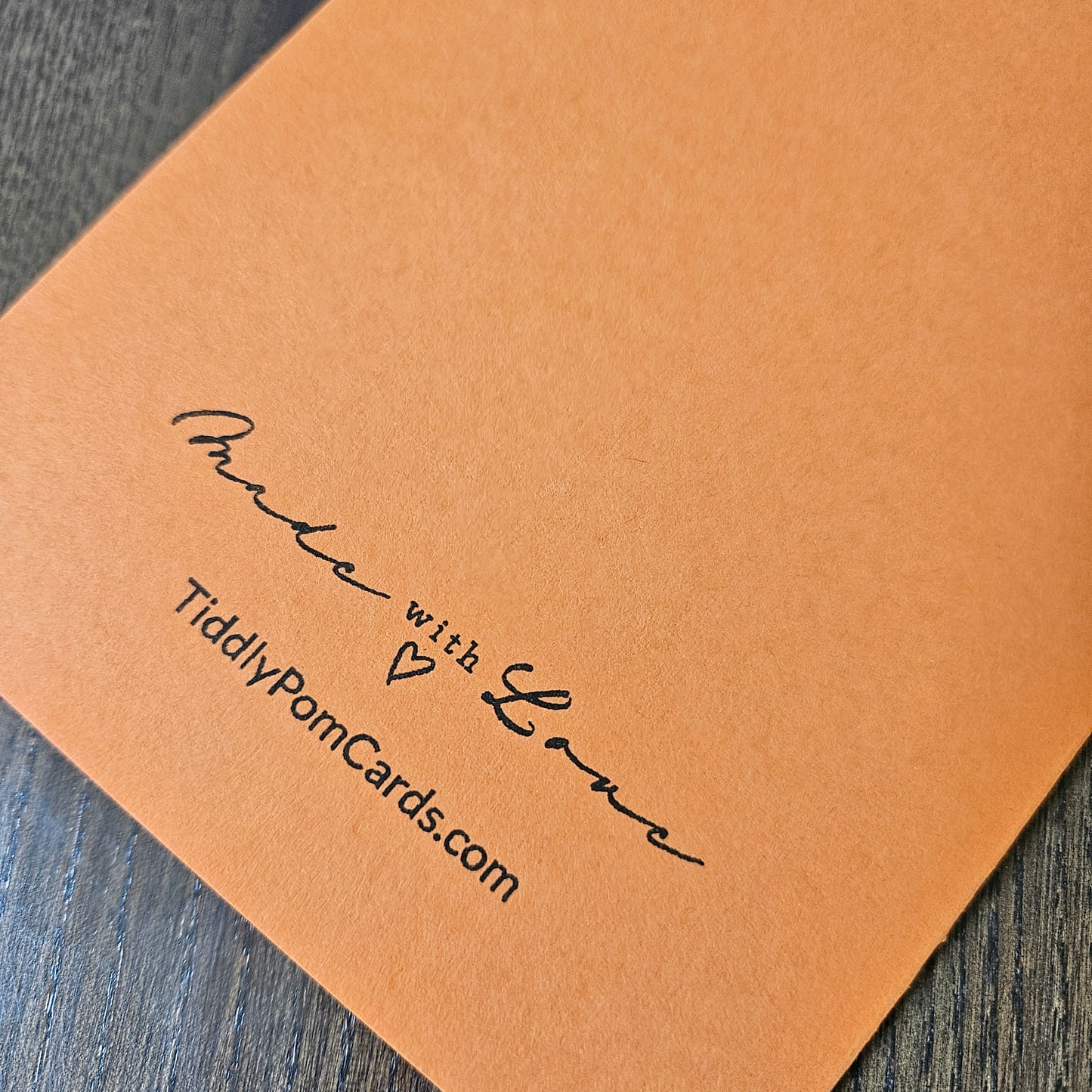 A close-up of a square tan pom card with black printed text reading 'Made with Love' and 'TiddlyPomCards.com', placed on a dark wood surface.