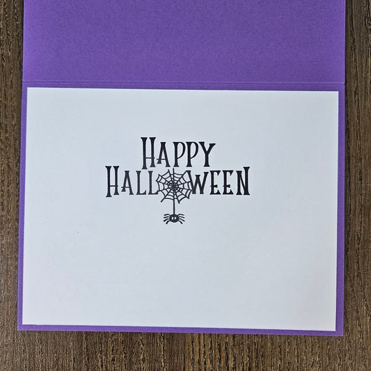 A square purple greeting card with a white front insert printed with 'Happy Halloween' in black decorative lettering and a spiderweb motif, photographed on a wood surface.