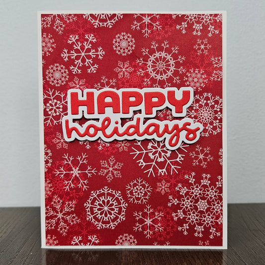 A handmade holiday greeting card with a red background, white snowflake patterns, and a layered 'HAPPY holidays' text design, standing on a dark wooden surface against a light gray wall.