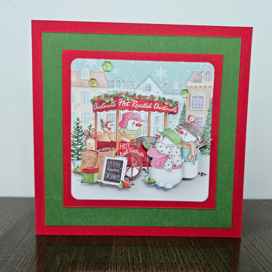 A handmade layered Christmas greeting card featuring a festive winter scene with snowman characters, a hot dog stand, and holiday decorations. The card has a red outer border, a green inner border, and a white central panel with a detailed illustration.