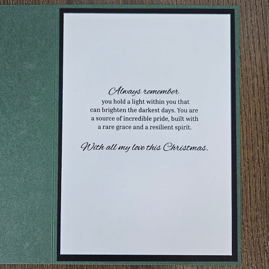 A square Christmas greeting card with a white center panel featuring a black script poem, set against a textured dark green cover, displayed on a brown wooden surface.