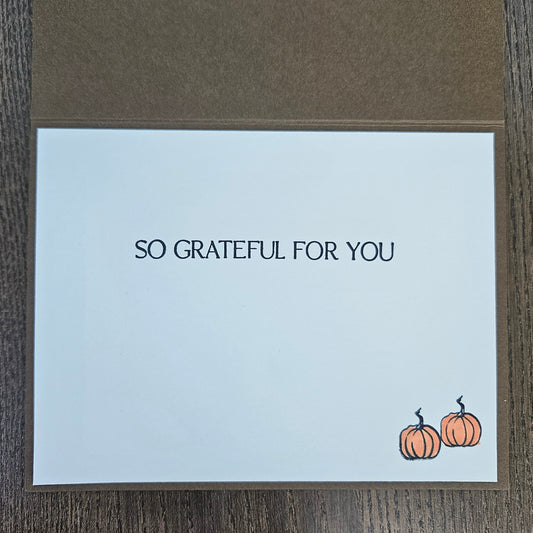 A square greeting card with a white front and brown kraft envelope, placed on a dark wood surface. The card reads 'SO GRATEFUL FOR YOU' in black text, with two small orange pumpkins near the bottom right corner.