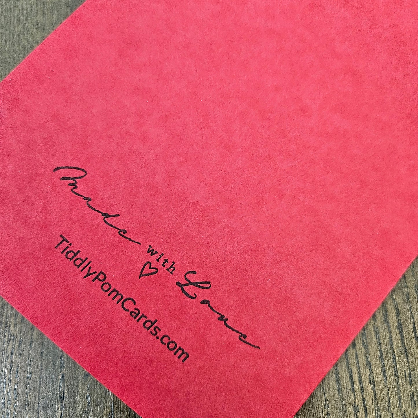 A close-up of a coral pink textured cardstock or paper sheet with black printed script reading 'Made with Love' and 'TiddlyPomCards.com', placed on a gray wood surface.
