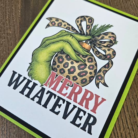 A square greeting card featuring a green hand holding a leopard-print ornament with a leopard-print bow, above the text 'MERRY WHATEVER' in red and black, photographed on a wood surface.