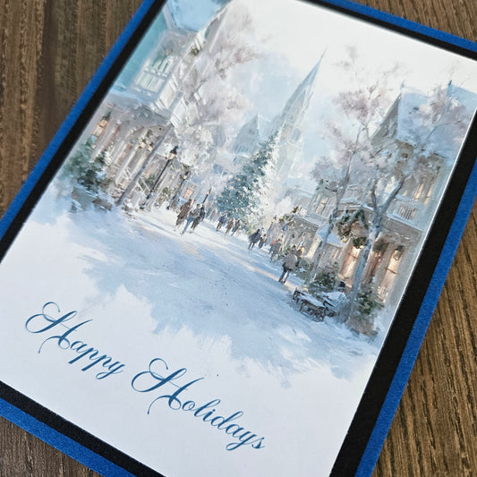 A square holiday greeting card featuring a watercolor winter street scene with snow-covered buildings, trees, and people, with the words 'Happy Holidays' in blue script at the bottom. The card is photographed at an angle on a wooden surface, bordered by a blue and black frame.