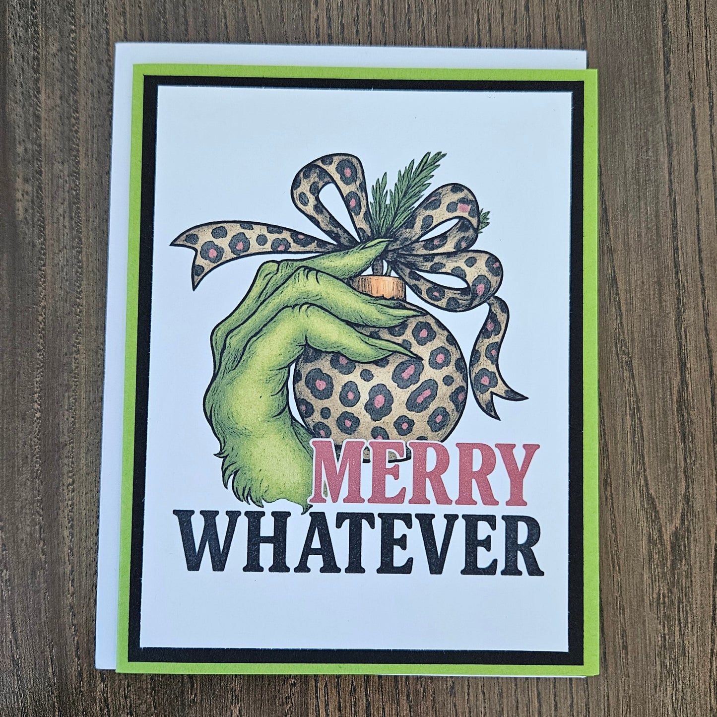 A square greeting card with a white background and a bright lime green border, featuring a hand holding a leopard-print ornament with the text 'MERRY WHATEVER' on a wooden tabletop.