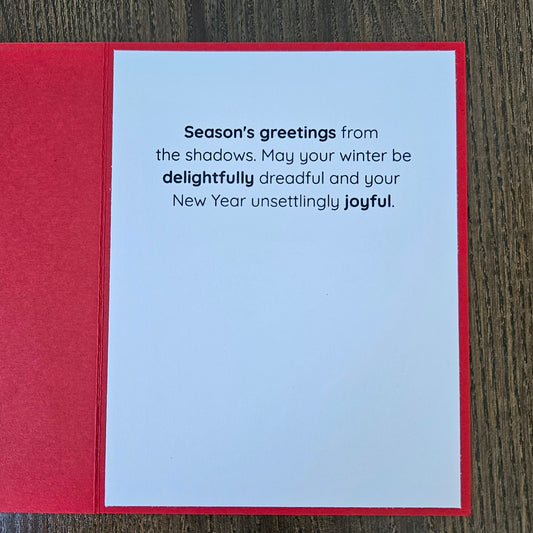 A red greeting card laid flat on a dark wood surface, showing a white interior with printed New Year's message: 'Season's greetings from the shadows. May your winter be delightfully dreadful and your New Year unsettlingly joyful.
