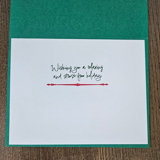 A square white greeting card with a green textured backing, placed on a brown wood surface. The front of the card reads, 'Wishing you a relaxing and stress-free holiday' in black script, with a small red decorative line and dots underneath.