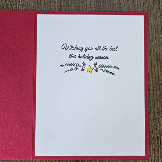 A handmade holiday greeting card with a white front insert and a textured red backing, placed on a dark wood background. The card features the printed text 'Wishing you all the best this holiday season.