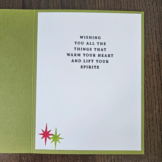 A square greeting card with a white front and olive green backing, placed on a dark wood surface. The front has centered black text reading, 'WISHING YOU ALL THE THINGS THAT WARM YOUR HEART AND LIFT YOUR SPIRITS,' with two small starburst motifs in red and green near the lower left corner.