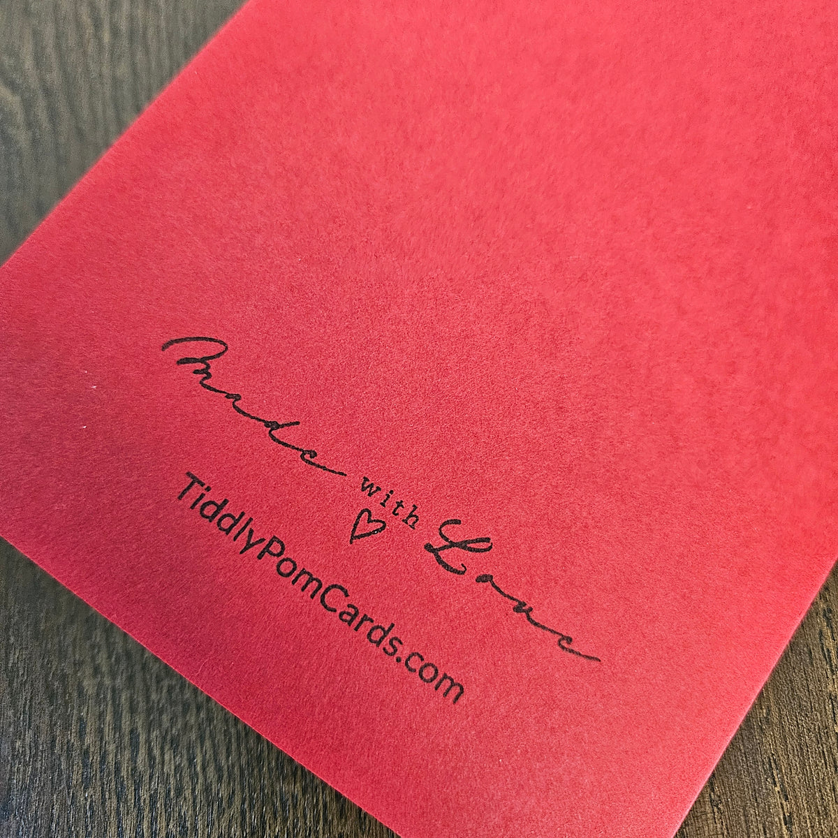 A close-up of a textured red card or envelope corner with black printed text reading 'Made with Love' and 'TidilyPomCards.com' on a dark wood surface.