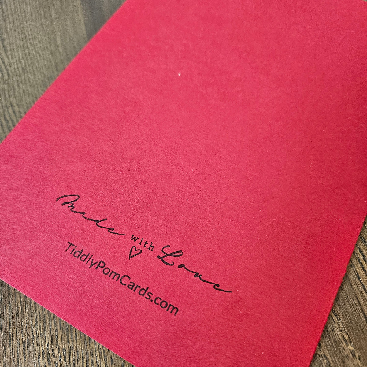 A close-up of a bright red textured card or envelope corner printed with the words 'Made with Love' and 'TidilyPomCards.com' in black script, placed on a wood surface.