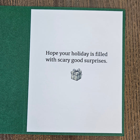 A greeting card with a dark green textured cover and a white insert card that reads, 'Hope your holiday is filled with scary good surprises.' above a small gift box illustration, photographed on a wood surface.