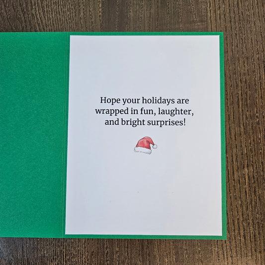 Printed inside of a green Christmas card reading Hope your holidays are wrapped in fun, laughter, and bright surprises, with a small Santa hat below.